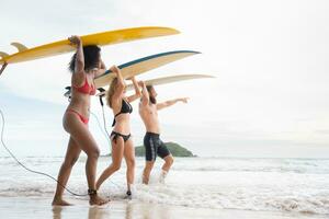 posteriore Visualizza di Due donne e giovane uomo Tenere tavole da surf su loro teste. e camminare in il mare per Surf foto