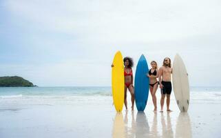 gruppo di amici nel costumi da bagno in posa con tavole da surf su il spiaggia. foto