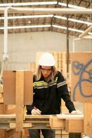 femmina falegname Lavorando nel un' la lavorazione del legno fabbrica, lei è indossare un' sicurezza casco e occhiali. controllo il pulizia di il tavole contro il design documenti. foto