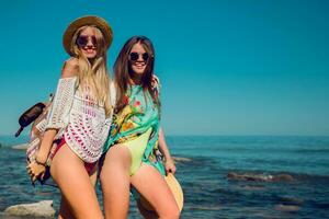 Due bellissimo amici a piedi nel il spiaggia e avendo divertimento. ragazze indossare elegante costumi da bagno , cannuccia cappello e freddo occhiali da sole. estate luminosa colori. foto