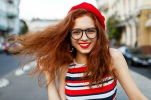 elegante giovane Zenzero donna nel rosso berretto e bicchieri in posa su il strada. foto