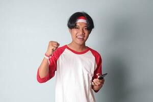 ritratto di attraente asiatico uomo nel maglietta con rosso e bianca nastro su testa, Tenere mobile Telefono mentre raccolta il suo cazzotto, festeggiare successo. isolato Immagine su grigio sfondo foto
