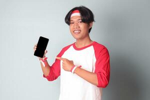 ritratto di attraente asiatico uomo nel maglietta con rosso e bianca nastro su testa, Tenere e mostrando vuoto schermo di mobile Telefono per modello. isolato Immagine su grigio sfondo foto