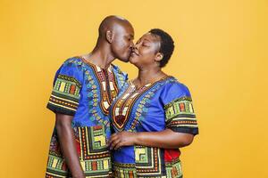 uomo baci sorridente allegro moglie nel guancia e mostrando amore. africano americano romantico coppia con contento espressione indossare etnico Abiti in posa nel studio su arancia sfondo foto