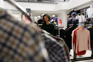 infelice insoddisfatto asiatico donna cliente in piedi nel capi di abbigliamento memorizzare guardare attraverso grucce con Abiti. femmina acquirente sensazione deluso con nero Venerdì i saldi mentre shopping nel moda centro commerciale foto