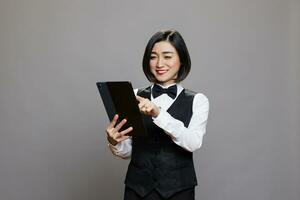 sorridente ristorante addetto alla reception nel uniforme controllo ospite elenco su digitale tavoletta. ristorante asiatico donna cameriera maschiatura su touch screen mentre l'esame in linea ordine mentre in posa nel studio foto