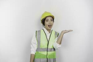 un' scioccato asiatico donna lavoro duro e faticoso indossare sicurezza casco e veste, puntamento per copia spazio accanto suo, isolato di bianca sfondo. lavoro giorno concetto. foto