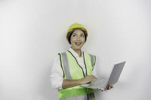 un' riflessivo giovane donna lavoro duro e faticoso lavoratore indossare sicurezza casco e veste mentre Tenere sua il computer portatile e guardare a parte per copia spazio, isolato di bianca sfondo. lavoro giorno concetto. foto