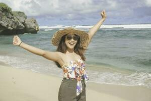 un' contento giovane asiatico donna indossare spiaggia cappello è in posa per il telecamera con sua mani aprire, esprimendo la libertà e spensierato vacanza a il spiaggia nel gunungkidul, Indonesia foto