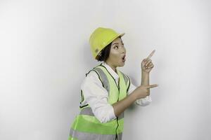 un' scioccato asiatico donna lavoro duro e faticoso indossare sicurezza casco e veste, puntamento per copia spazio accanto suo, isolato di bianca sfondo. lavoro giorno concetto. foto