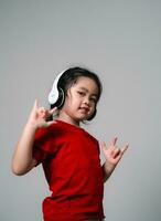 allegro contento asiatico bambini ragazza con nero capelli ascoltando per musica canzoni nel cuffie su un' grigio sfondo. ritratto carino ragazza attenzione rosso camicia e guardare a telecamera studio illuminazione. foto