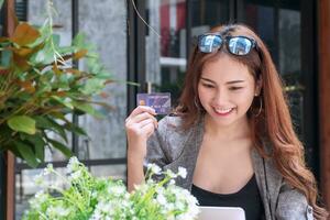 sorridente giovane donna con il computer portatile shopping in linea Tenere credito carta con occhiali da sole seduta nel giardino sfondo. attraente attività commerciale donna ottenere soddisfare e contento con la libertà di pagamento. foto