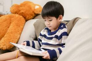 asiatico ragazzo lettura un' libro su il divano apprendimento al di fuori il aula foto