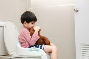 asiatico ragazzo seduta su il gabinetto ciotola nel mano Tenere orsacchiotto orso foto