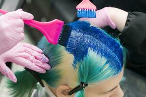 Due parrucchieri utilizzando rosa spazzola mentre l'applicazione blu dipingere per femmina con Smeraldo capelli colore durante processi di tintura capelli nel unico lapis lazuli colore foto