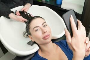 brunetta donna sembra a smartphone mentre parrucchiere è occupato lavaggio sua capelli a bellezza salone foto