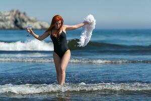 donna nel costume da bagno a piedi caviglia in profondità in onde di oceano. femmina Tenere vino bicchiere e boa nel mani foto