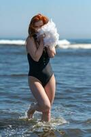 sexy donna nel costume da bagno a piedi nel onde di oceano, Tenere boa nel mani e scherzosamente copertura viso foto