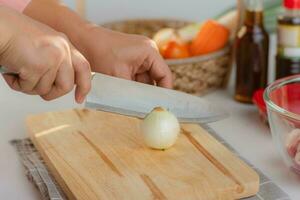 donna mano utilizzando coltello per tagliare bianca cipolla su di legno taglio tavola. donna preparazione cibo nel il cucina a casa. foto