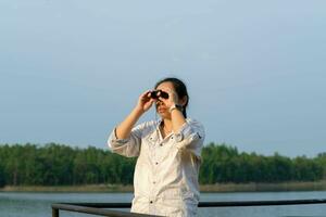 giovane femmina esploratore con binocolo esplorando natura o Guardando uccelli all'aperto. giovane donna guardare attraverso binocolo a uccelli su il serbatoio. osservazione uccelli foto