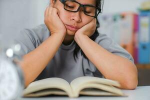 stanco asiatico donna seduta a il scrivania lettura un' libro nel il vivente camera. foto