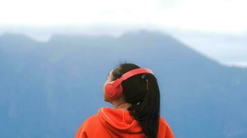 posteriore Visualizza di un' giovane donna in piedi su un' calma cima e ascoltando per musica nel cuffie nel il mattina. donna indossare un' maglione godendo il bellezza di natura guardare a il montagna nel inverno. foto