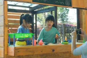 Due contento sorelle giocare con cibo e di legno drogheria giocattoli a il all'aperto terreno di gioco con sua madre. carino ragazze gioco di ruolo vendita frutta succo a il parco. famiglia la spesa tempo insieme su vacanza. foto