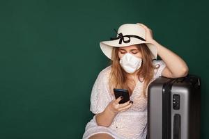 ragazza turista in una maschera medica si siede con i bagagli su uno sfondo verde