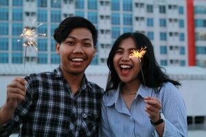 contento asiatico coppia Tenere sparkler guardare a telecamera con sorridente viso, celebrare nuovo anni vigilia, su urbano edificio sfondo, indossare casuale camicia nel all'aperto foto