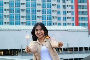 contento asiatico giovane donna fricchettone Tenere sparklers fuoco d'artificio celebrare nuovo anno vigilia con danza nel tetto superiore appartamento con urbano edificio sfondo foto