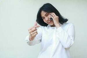 ritratto di spensierato asiatico donna giovane imprenditore mostrando coreano amore segni per telecamera mentre A proposito di su mobile Telefono indossare bianca formale camicia, in piedi isolato sfondo foto