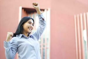 vicino su di sorridente asiatico donna d'affari indossare formale completo da uomo festeggiare vincente vittoria in piedi al di sopra di urbano costruzione, femmina imprenditore raccolta cazzotto dire sì gesto in piedi all'aperto foto