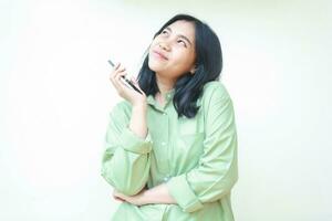 felice spensierato asiatico donna guardare su per vuoto spazio mentre parlando su smartphone hold su mani indossare verde oversize camicia isolato su bianca foto