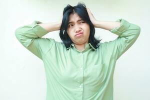 stressato asiatico donna con buio capelli afferrando sua testa guardare lontano confuso indossare verde sovradimensionato camicia, frustrato femmina sentire sotto pressione, isoated su bianca sfondo foto