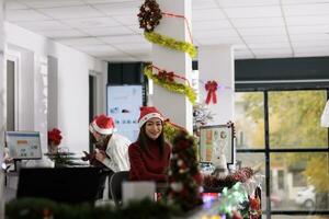 ritratto di gioioso asiatico donna d'affari Lavorando su Natale stagione nel decorato ufficio. sorridente femmina dipendente soluzione compito a scrivania nel diverso posto di lavoro durante inverno festivo tempo foto