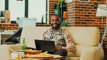 sorridente uomo utilizzando il computer portatile nel vivente camera e Guardando tv, sensazione rilassato con multiplo consegna pasti e birra. giovane tipo navigazione Internet su pc, seduta nel davanti di televisione. tripode sparo. foto