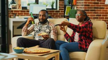 africano americano partner mangiare Pizza fette a casa, sensazione contento Guardando film insieme. rilassato coppia avendo divertimento mangiare porta via consegna cibo nel vivente camera, tempo libero attività. foto