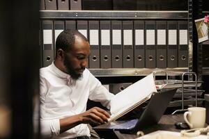 africano americano accusatore lettura file, preparazione per Astuccio nel polizia ufficio con Archivio storico record. legge rinforzo professionale analizzando legale competenza cartella a notte tempo foto