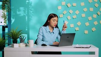 annoiato dipendente Lavorando noioso monotono poco brillante noioso ufficio lavoro. asiatico donna d'affari giocando con penna mentre Lavorando su il computer portatile nel triste poco interessante posto di lavoro al di sopra di blu studio sfondo foto