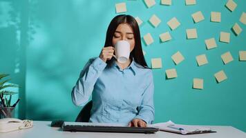 asiatico donna d'affari avendo tazza di caffè mentre nel in linea video conferenza incontro, accuratamente ascoltando e chat utilizzando tastiera del computer. dipendente nel moderno ufficio al di sopra di blu studio sfondo foto