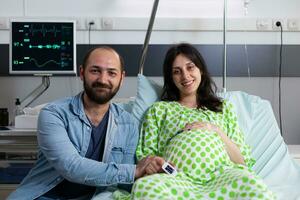 sorridente futuro genitori aspettando bambino nel ospedale reparto, preparazione per cesareo chirurgia. incinta donna guardare a telecamera mentre dire bugie nel letto in attesa per medico nel maternità clinica. parto concetto foto