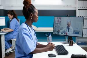 medico squadra su Internet video conferenza telemedicina video chiamata nel occupato clinico ufficio. africano americano infermiera a ospedale scrivania consulenza con generale professionista di paziente Astuccio foto