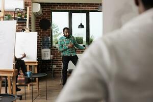 giovane africano americano uomo insegnamento arte per persone nel creativo studio, maschio istruttore dimostrando schizzi tecniche per studenti seduta a cavalletti durante gruppo disegno laboratorio foto