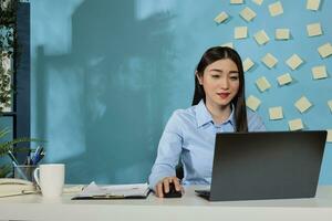 ritratto di un asiatico femmina dipendente Lavorando su il computer portatile per creare mensile rapporto. uomo d'affari seduta nel davanti di il computer nel il azienda spazio di lavoro con forniture su scrivania. foto