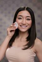 cosmetologia sorridente asiatico modello fare cura della pelle routine ritratto. allegro giovane bellissimo donna mostrando cura della pelle detergente passo utilizzando micellare acqua e guardare a telecamera foto