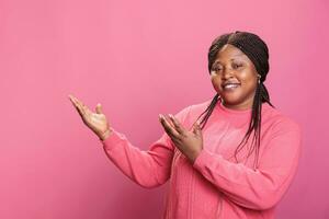 africano americano promotore puntamento per lato con palma mentre pubblicità Prodotto nel studio. allegro giovane adut mostrando promozione in piedi al di sopra di rosa sfondo, pubblicizzare gesto foto