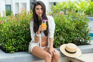 contento giovane bellissimo concia donna nel elegante boho bianca beachwear seduta vicino tropicale piscina nel lusso Hotel e godendo arancia cocktail o succo. spiaggia festa. foto