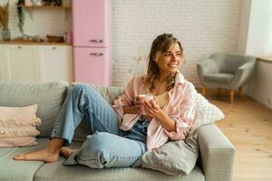 grazioso donna nel casuale Abiti riposo un divano nel accogliente vivente camera. Tenere tazza di tè. foto