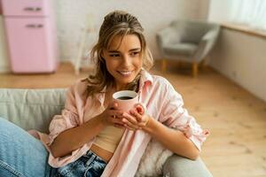 grazioso donna nel casuale Abiti riposo un divano nel accogliente vivente camera. Tenere tazza di tè. foto