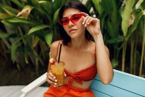 sbalorditivo abbronzatura donna nel retrò occhiali da sole rilassante nel tropicale spiaggia club nel elegante rosso superiore e arancia corti. potabile gustoso limonata. vacanza e vacanze. foto
